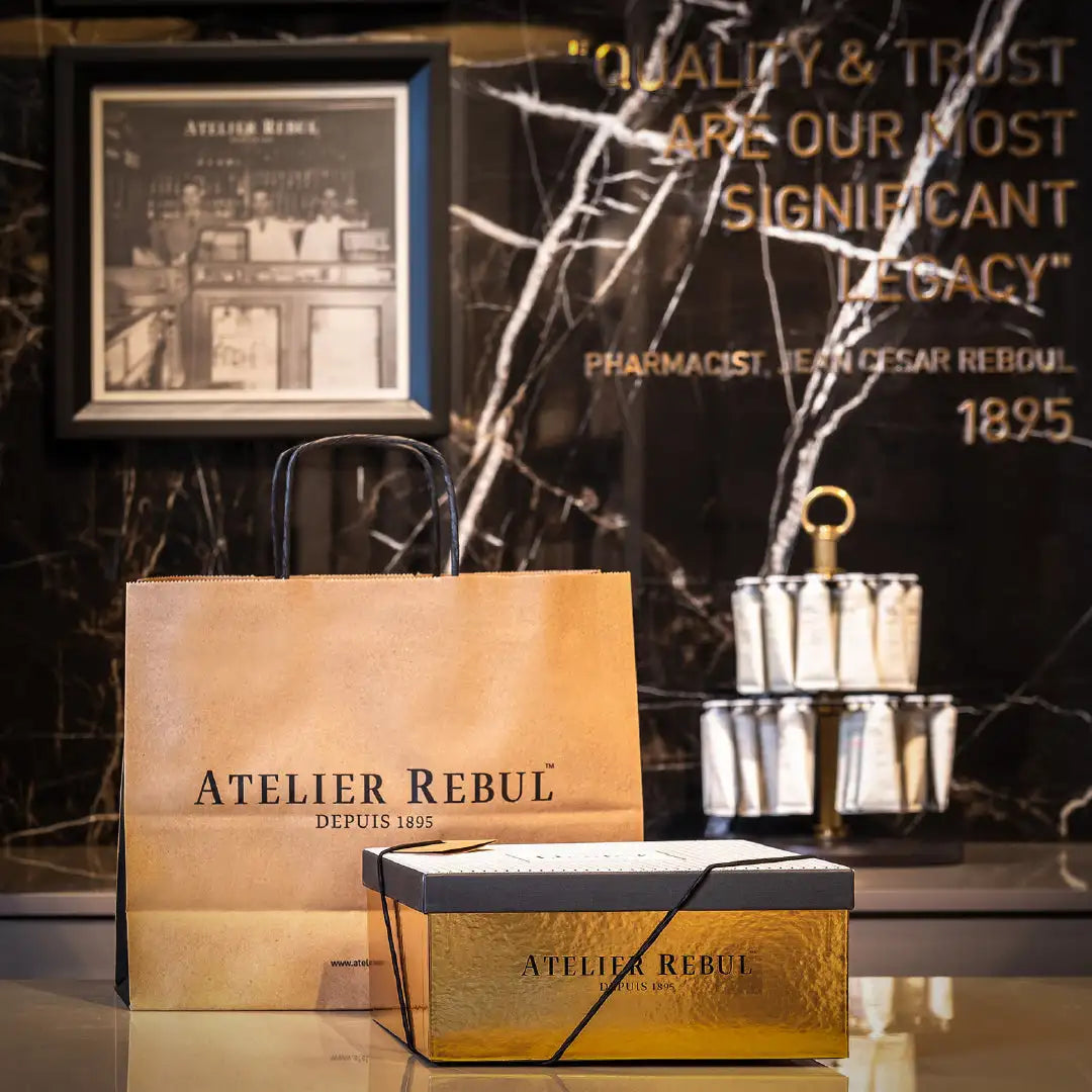 Cutie cadou dreptunghiulară căptușită cu folie de aur, cu panglică neagră și inscripția „atelier rebul depuis 1895” tipărită cu litere închise la culoare.
