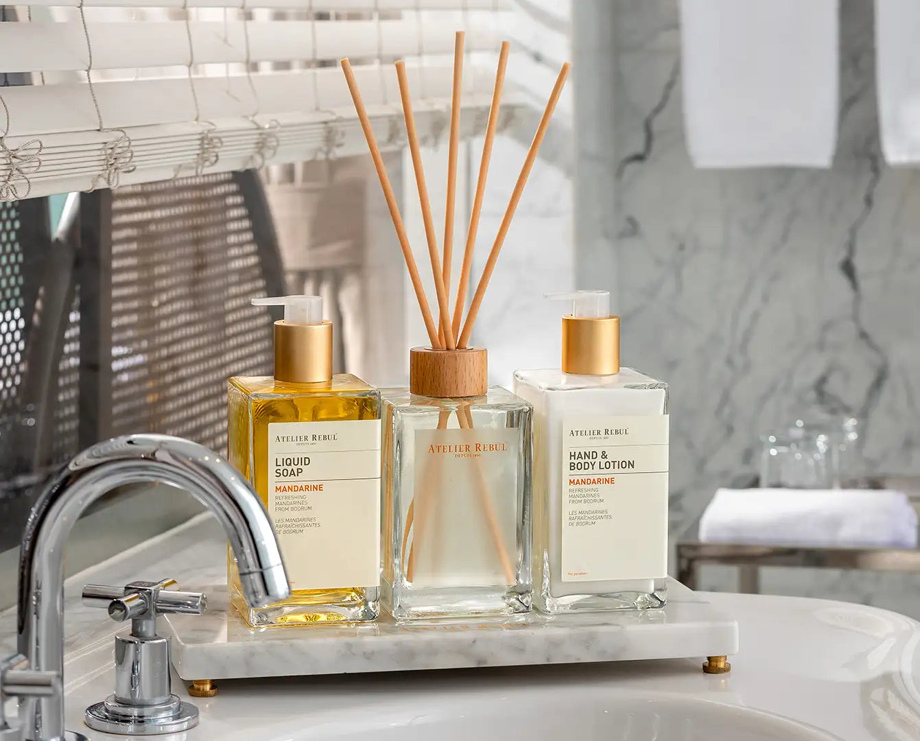 Difuzor de sticlă transparentă cu bețe de reed din lemn și un capac cu ton auriu, cu numele mărcii atelier rebe.