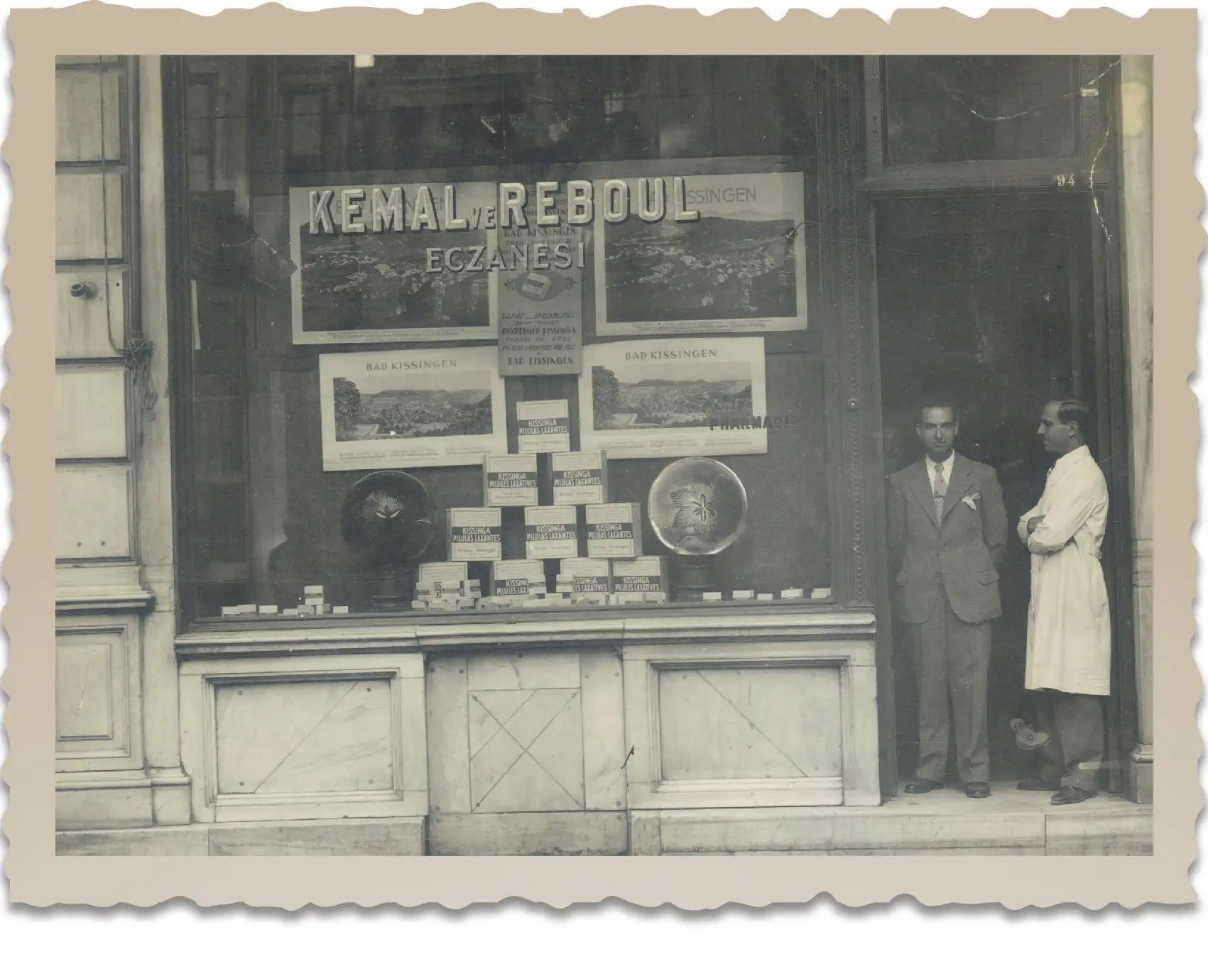 O fotografie vintage alb-negru a unei vitrine de magazin pentru ’kemal ve reboul eczanesi̇,’ cu cutii stivuite, postere cu imagini de peisaj și text, și doi bărbați stând în afara.