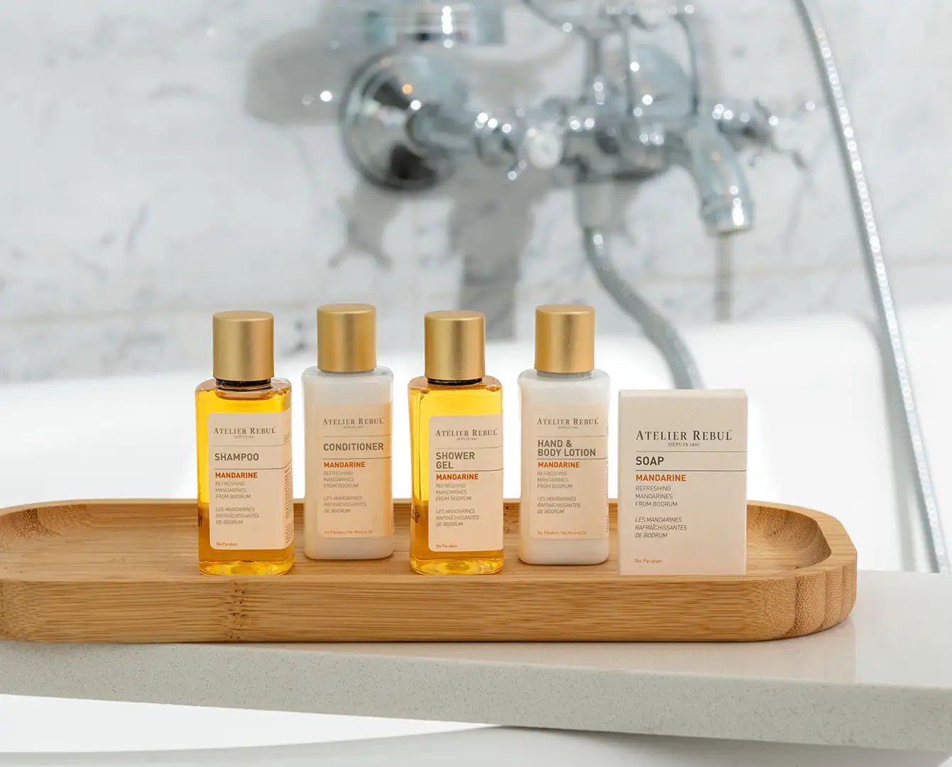 Set de cinci articole de toaletă de călătorie Atelier Rebul în sticle de sticlă amber cu capace aurii și un săpun solid bej, toate aranjate pe o tavă de lemn.