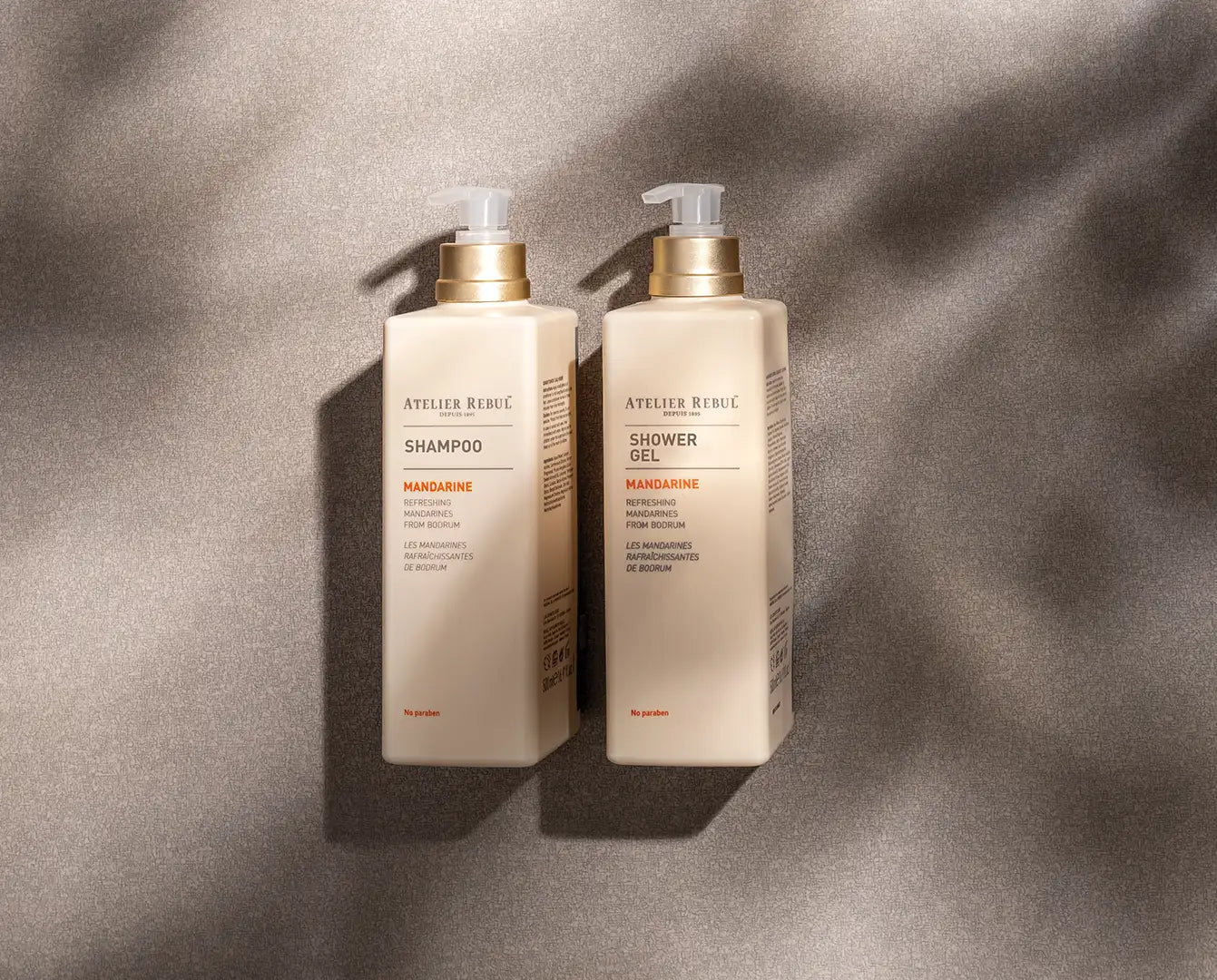 Sticle dreptunghiulare de culoare crem cu dozatoare cu pompă din aur, etichetate „atelier rebu” și cu un parfum de mandarină.